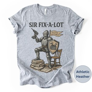 Peut inclure: T-shirt gris chin&eacute; avec l'inscription "SIR FIX-A-LOT" au-dessus d'une illustration d'un chevalier tenant une perceuse. Le chevalier est debout sur des manuels, avec un bouclier et une banni&egrave;re "DUCTUS MAXIMUS". Une petite chaise en bois est &agrave; droite.