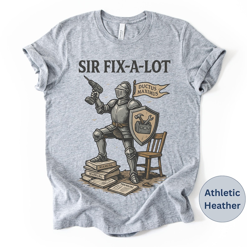 Peut inclure: T-shirt gris chin&eacute; avec l'inscription "SIR FIX-A-LOT" au-dessus d'une illustration d'un chevalier tenant une perceuse. Le chevalier est debout sur des manuels, avec un bouclier et une banni&egrave;re "DUCTUS MAXIMUS". Une petite chaise en bois est &agrave; droite.