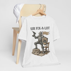 Peut inclure: T-shirt gris clair avec un chevalier de dessin anim&eacute; tenant une perceuse, avec le texte "SIR FIX-A-LOT" au-dessus. Le chevalier est debout sur des livres et porte un bouclier avec un marteau et une cl&eacute;. Une petite chaise en bois est &agrave; c&ocirc;t&eacute; du chevalier.