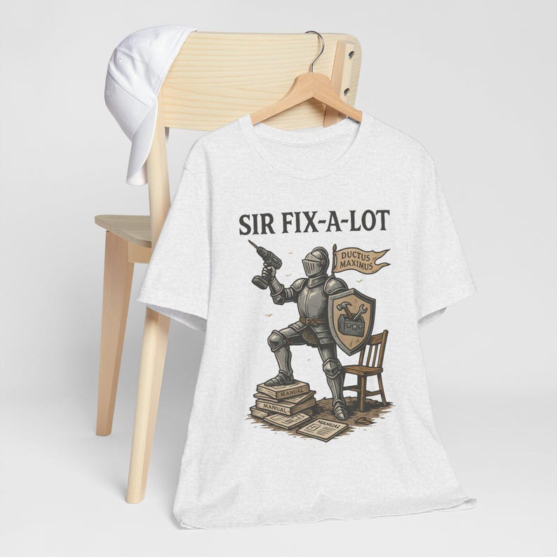 Peut inclure: T-shirt gris clair avec un chevalier de dessin anim&eacute; tenant une perceuse, avec le texte "SIR FIX-A-LOT" au-dessus. Le chevalier est debout sur des livres et porte un bouclier avec un marteau et une cl&eacute;. Une petite chaise en bois est &agrave; c&ocirc;t&eacute; du chevalier.