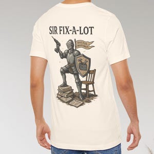 Peut inclure: T-shirt cr&egrave;me avec un chevalier de dessin anim&eacute; tenant une perceuse et un bouclier, avec le texte "SIR FIX-A-LOT". Le chevalier est debout sur des manuels, avec une petite chaise en bois. La banni&egrave;re dit "DUCTUS MAXIMUS".