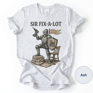 Peut inclure: T-shirt gris clair avec l'inscription "SIR FIX-A-LOT" et un chevalier en armure tenant une perceuse. Le chevalier est debout sur des manuels, avec un bouclier et une banni&egrave;re indiquant "DUCTUS MAXIMUS". Une petite chaise en bois est &agrave; droite.