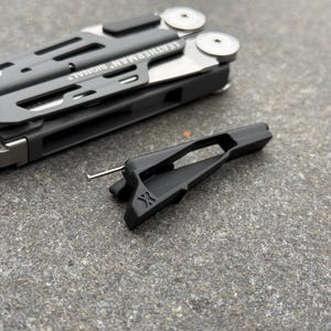 以下が含まれることがあります： 「LEATHERMAN SIGNAL」の文字が入った、ダークグレーとシルバーのマルチツール。黒いアクセサリーには「X」の文字が。グレーの表面に置かれた写真。