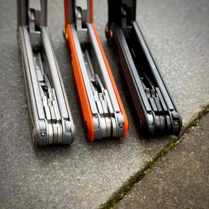 May include: Three multi-tools in a row, each with a different color scheme. The first is a light gray with silver tools, the second is orange with silver tools, and the third is black with silver tools. The tools are open, revealing various implements.