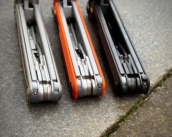 May include: Three multi-tools in a row, each with a different color scheme. The first is a light gray with silver tools, the second is orange with silver tools, and the third is black with silver tools. The tools are open, revealing various implements.