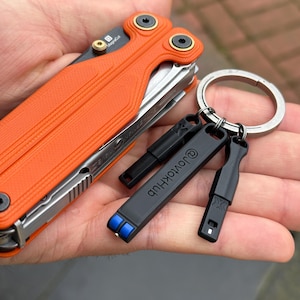 Op de afbeelding: Een multitool met feloranje getextureerde schalen en zwarte hardware, voorzien van een MagnaCut-bladmarkering. Aan een zilverkleurige metalen sleutelhanger hangen drie kleine zwarte accessoires, waarvan er één is gegraveerd met de tekst @JovtokHub.