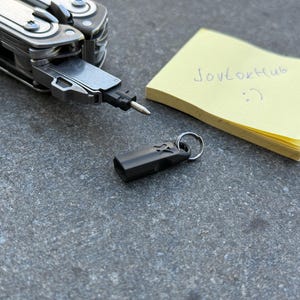 Op de afbeelding: Een multitool met een zwart opzetstuk en een kleine, zwarte sleutelhanger. Er ligt ook een gele plaknotitie met de tekst "Jovtokhub :) ". De items liggen op een grijs oppervlak.