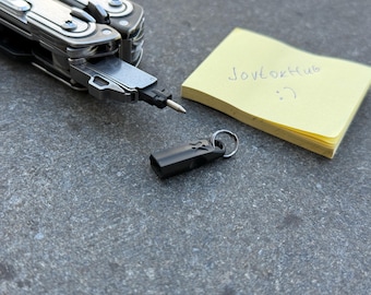 Puede incluir: Una multiherramienta con un accesorio negro y un pequeño llavero negro. También hay una nota adhesiva amarilla con el texto "Jovtokhub :) ". Los objetos están sobre una superficie gris.