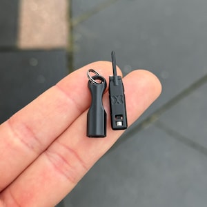 Puede incluir: Dos pequeñas herramientas negras. Una es un destornillador con punta y la otra es una pequeña herramienta cilíndrica con un llavero. El destornillador tiene la letra "X" impresa. Ambas herramientas se sostienen en una mano.