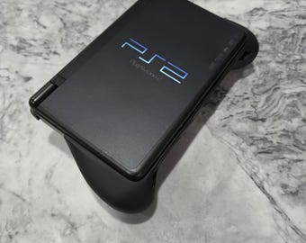 May include: A black handheld gaming console featuring a matte finish and a hinged design. The top surface displays the blue PS2 logo and the text PlayStation 2. The device is integrated into a black ergonomic grip controller attachment.