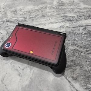 May include: A handheld gaming console featuring a matte black frame with a deep red top cover. The cover displays a circular blue lens, three small yellow indicator lights, and a small yellow triangle icon. The device is fitted with black ergonomic grips.