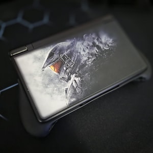 Puede incluir: Una consola de juegos portátil negra con una funda protectora. La pantalla muestra un gráfico detallado de un casco de soldado futurista con una visera naranja. El dispositivo descansa sobre una superficie negra.