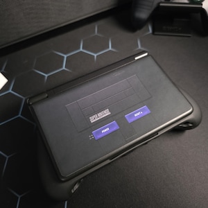 May include: A black handheld gaming console with the Super Nintendo logo and purple buttons labeled "Power" and "Reset". The console is on a black surface with a hexagonal pattern.