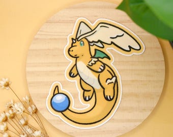 Könnte beinhalten: Ein gelber Aufkleber mit einem Cartoon-Drachen mit weißen Flügeln und einer blauen Kugel am Schwanz. Der Drache hat einen weißen Hut und grüne Akzente. Der Aufkleber befindet sich auf einer Holzoberfläche.