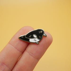 Könnte beinhalten: Ein schwarz-weißer Emaille-Ansteckpin in Form eines Vogels. Der Vogel hat eine silberne Umrandung und einen kleinen, gebogenen Schnabel. Der Pin liegt auf einem hellorangen Hintergrund.