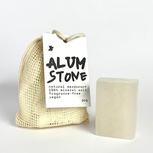 May include: A natural deodorant alum stone with a tag that reads "ALUM STONE". The tag also states the product is a natural deodorant, 100% mineral salt, fragrance-free, and vegan. The alum stone is next to a small beige mesh bag.