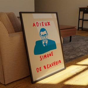 May include: A framed poster with a black frame and a cream background. The poster features a blue illustration of a person with glasses and the text "Adieu Simone de Beauvoir" in red.