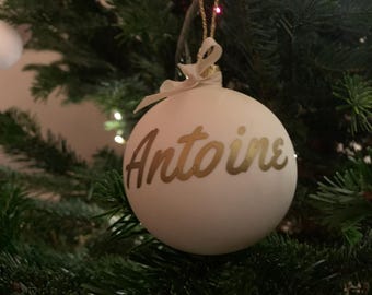 Könnte beinhalten: Eine weiße Weihnachtskugel mit dem Namen "Antoine" in goldener Schrift. Der Schmuck hängt an einer goldenen Schnur und ist mit einer weißen Schleife gebunden. Der Schmuck wird an einem grünen Weihnachtsbaum ausgestellt.