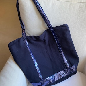 Peut inclure: Sac fourre-tout bleu marine avec des détails de sequins. Le sac a une forme rectangulaire avec de longues anses et une large ouverture. Les anses et le bas sont ornés de sequins bleus scintillants.