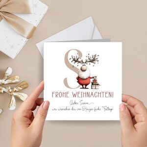 Pu&ograve; includere: Biglietto di Natale con un personaggio renna in abito rosso, con regali. Il biglietto presenta il testo "FROHE WEIHNACHTEN!" e un messaggio scritto a mano. Lo sfondo &egrave; beige chiaro.