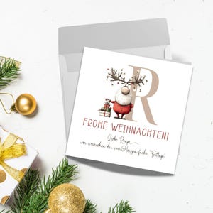 Pu&ograve; includere: Un biglietto di Natale con un design festivo con un ibrido renna-Babbo Natale che tiene dei regali. La carta ha la lettera "R" e le parole "Frohe Weihnachten!" in rosso. Una busta e decorazioni natalizie sono sullo sfondo.