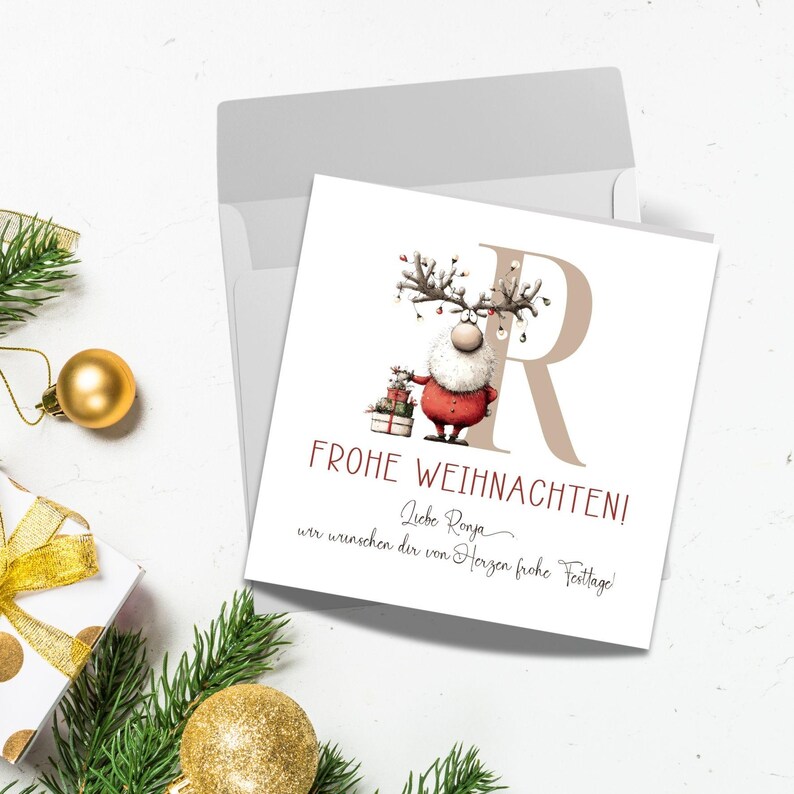 Pu&ograve; includere: Un biglietto di Natale con un design festivo con un ibrido renna-Babbo Natale che tiene dei regali. La carta ha la lettera "R" e le parole "Frohe Weihnachten!" in rosso. Una busta e decorazioni natalizie sono sullo sfondo.