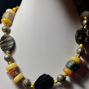 May include: A handcrafted necklace featuring a variety of beads in shades of yellow, black, silver, and earth tones. The necklace includes round, oval, and irregularly shaped beads, with a prominent black shell-shaped pendant. The beads are strung together with yellow string.