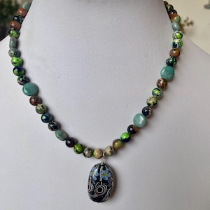 Può includere: Collana di perline con pendente. La collana presenta perline rotonde verdi, marroni e blu. Il pendente ovale ha uno sfondo nero e un motivo floreale.