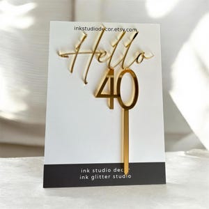 May include: A gold-coloured cake topper with the word "Hello" in cursive and the number "40". The topper is attached to a long, thin stem. The background is a white card with the text "inkstudio decor.etsy.com".