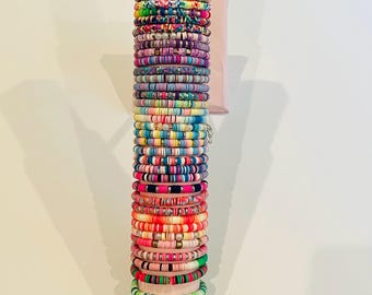 Puede incluir: Una pila vertical de numerosos brazaletes de cuentas de arcilla de colores dispuestos en un expositor cilíndrico. Los brazaletes presentan varios patrones, incluyendo colores sólidos, rayas y acentos metálicos. Una pulsera superior muestra la palabra SUMMER.