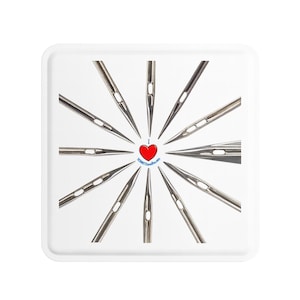 Op de afbeelding: Een witte vierkante tray met een radiale opstelling van naainaalden. In het midden een rood hart met de tekst "I ❤️ SCHMETZ needles.com" in een blauwe cirkel. De naalden zijn zilverkleurig en wijzen naar buiten.