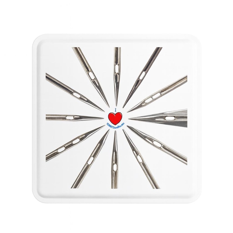 Op de afbeelding: Een witte vierkante tray met een radiale opstelling van naainaalden. In het midden een rood hart met de tekst "I ❤️ SCHMETZ needles.com" in een blauwe cirkel. De naalden zijn zilverkleurig en wijzen naar buiten.