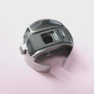 May include: A close-up of a silver sewing machine bobbin case. The metal bobbin case has a rectangular opening and a curved base. The background is a pale pink colour.