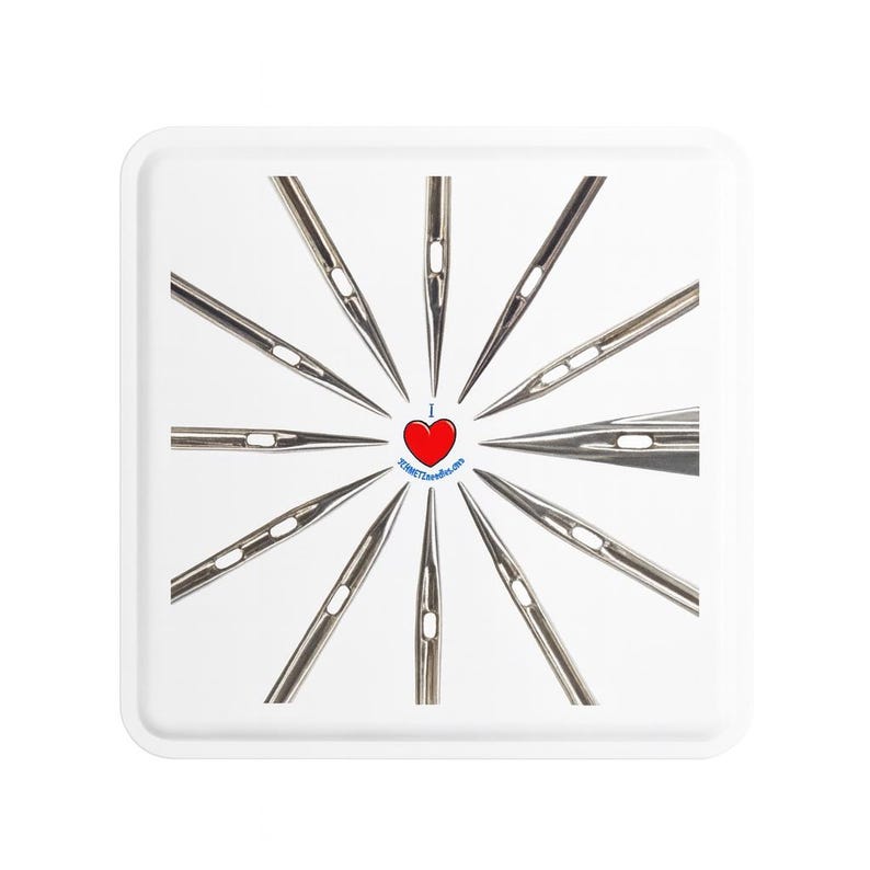 Op de afbeelding: Een witte vierkante tray met een radiale opstelling van naainaalden. In het midden een rood hart met de tekst "I ❤️ SCHMETZ needles.com" in een blauwe cirkel. De naalden zijn zilverkleurig en wijzen naar buiten.