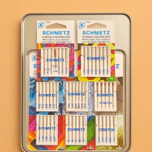 May include: A metal tin containing multiple packs of SCHMETZ sewing machine needles. The packs are labeled with the SCHMETZ logo and needle type, including "Costume Pack" and "Stretch" needles. The tin is on a light orange background.
