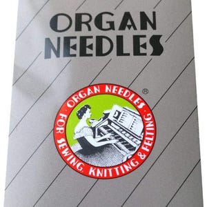 Può includere: Una confezione di aghi Organ, con sfondo argentato e linee diagonali grigie. Il testo "ORGAN NEEDLES" è in nero. Un emblema circolare mostra una donna che suona un organo. Il testo "FOR SEWING, KNITTING & FELTING" circonda l'emblema.