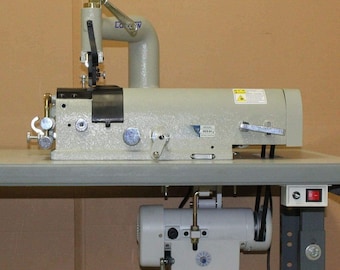 May include: A beige industrial sewing machine with a curved arm and a motor attached underneath. The machine has various levers, dials, and a silver-colored needle component. It is mounted on a gray table.