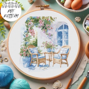 May include: A cross-stitch pattern of a patio scene with two wicker chairs, a small table, and a potted plant. The chairs are blue and white, and the table is brown. The potted plant is green and has pink flowers. The background is a white wall with a blue window.