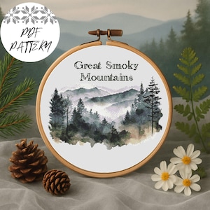 May include: Cross-stitch embroidery in a wooden hoop featuring a misty mountain landscape with the text "Great Smoky Mountains." The design includes trees and a soft color palette of greens, grays, and whites. A "PDF PATTERN" logo is in the upper left.