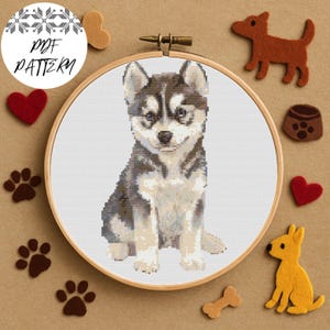 Può includere: Un ricamo a punto croce di un cucciolo di husky in un telaio di legno. Il cucciolo è grigio, nero e bianco. Le parole "PDF PATTERN" sono in un cerchio sopra il telaio. L'immagine è circondata da decorazioni in feltro a tema cani.