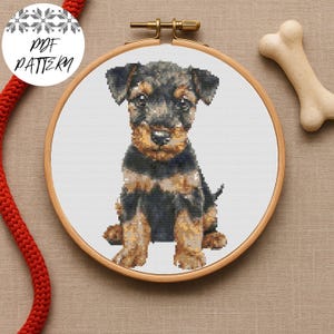 以下が含まれることがあります： 木製の刺繍枠に入ったテリアの子犬のクロスステッチパターン。子犬は黒とタンで、詳細なステッチが施されています。白い犬の骨と赤いロープも表示されています。「PDF PATTERN」という文字が白い円にあります。