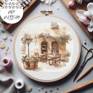 Puede incluir: Un patrón de punto de cruz de una escena de cafetería con un edificio, mesas, sillas y plantas. El patrón está en un aro de bordado redondo.