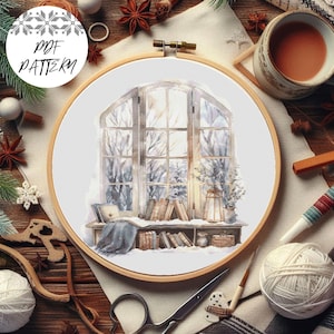 Puede incluir: Un patrón de punto de cruz que presenta una ventana con vista a una escena de invierno nevada. La ventana está enmarcada por un alféizar de madera con libros y una linterna. El patrón está en un aro blanco.