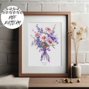 Puede incluir: Un patrón de punto de cruz enmarcado con un ramo de flores rosas, moradas y azules, atado con una cinta morada. El texto "September Aster" está debajo. El marco es marrón y el fondo es blanco.