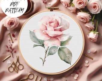 May include: A cross stitch pattern of a single pink rose with green leaves in a wooden embroidery hoop. The hoop is on a pink fabric background with other pink roses.