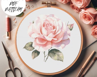 May include: A cross stitch pattern featuring a single pink rose with green leaves. The pattern is displayed in a wooden embroidery hoop on a white fabric background.