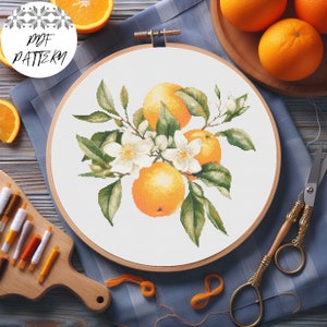 May include: A white embroidery hoop with a cross-stitch design of orange blossoms and oranges. The hoop is on a blue and white checkered fabric. There are orange slices and embroidery floss nearby.