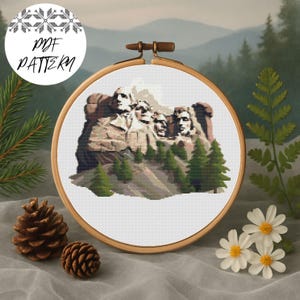 Puede incluir: Un patrón PDF de punto de cruz con el Monte Rushmore, enmarcado en un bastidor de bordado de madera. El diseño muestra el monumento con árboles verdes y un fondo de montaña. El texto "PDF PATTERN" está en un círculo blanco.
