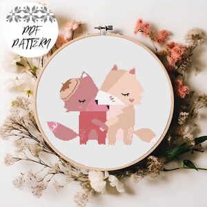 Könnte beinhalten: Kreuzstichmuster von zwei sich umarmenden Füchsen, einer trägt einen braunen Hut. Die Füchse sind rosa, braun und weiß. Das Muster ist in einem weißen Stickrahmen.