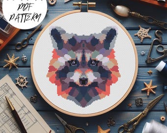 May include: A cross-stitch pattern of a raccoon's face in a wooden embroidery hoop. The design features geometric shapes in shades of purple, orange, and blue. The text "PDF PATTERN" is in a white circle in the upper left corner.